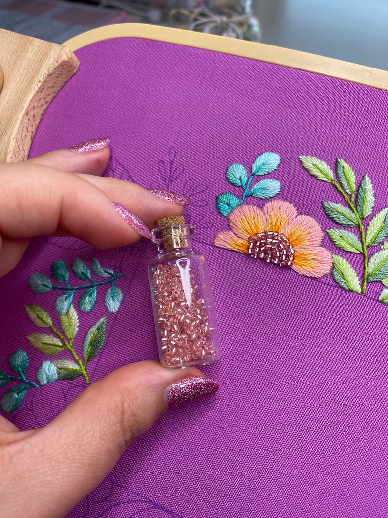 Op de afbeelding: Een kleine glazen fles gevuld met roze kralen, gehouden voor een paarse stof met geborduurde bloemen en bladeren. Het borduurwerk heeft tinten roze, oranje, groen en blauw. De fles heeft een kurk.