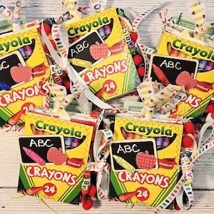 May include: Several handmade gift bags with a Crayola crayon box theme. Each bag features a yellow crayon box design with the words "Crayola", "Crayons", and "24". The bags are decorated with ribbons, small red pom-poms, and apple-shaped cutouts.