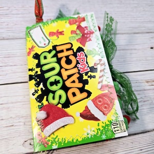 Könnte beinhalten: Eine grüne und gelbe Sour Patch Kids Süßigkeitenbox mit einem roten und weißen Weihnachtsmann-Hut, einem Zuckerstangen und einer grün-weißen Schleife. Die Schachtel trägt die Aufschrift "Sour Patch Kids" und "110 Kalorien pro 12 Stück".