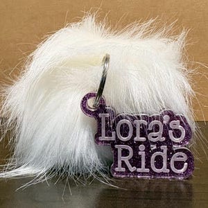 Puede incluir: Un llavero con un pompón blanco y esponjoso y una etiqueta con purpurina morada. La etiqueta dice "Lora's Ride" en letras blancas. El llavero está sujeto con un anillo de metal plateado.
