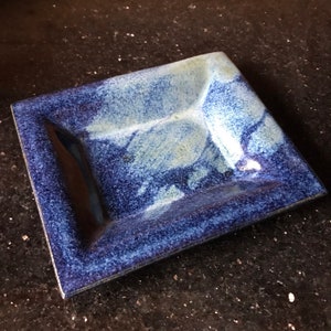 May include: A square ceramic dish with a blue and white glaze. The dish has a raised rim and a slightly textured surface.