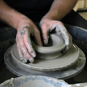 May include: A person's hands are shaping a lump of clay on a pottery wheel. The clay is being spun and moulded into a bowl shape.