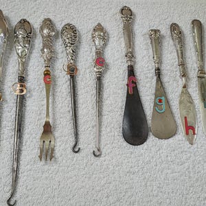 Peut inclure: Collection d'ustensiles de service anciens en argent, comprenant des fourchettes, des couteaux et des chausse-pieds. Chaque pièce présente des manches ornés et des motifs décoratifs. Certains ustensiles ont des lettres colorées.