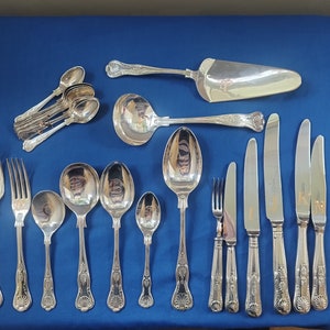 Puede incluir: Un conjunto de cubiertos de plata antiguos, que incluye tenedores, cucharas, cuchillos y utensilios para servir, dispuestos sobre una superficie azul. La cubertería presenta detalles ornamentales en los mangos y un acabado pulido. El juego incluye una paleta para tartas y un cucharón.