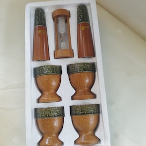 Może przedstawiać: Zestaw sześciu drewnianych stojaków na jajka z zielono-brązowymi wzorami. Zestaw zawiera również dwie drewniane solniczki i pieprzniczki oraz drewniany klepsydrę.