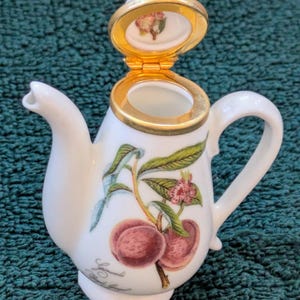 Puede incluir: Una tetera de porcelana blanca con tapa dorada y diseño de melocotón. La tapa está abierta y el interior de la tetera es visible.