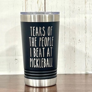 Può includere: Un bicchiere termico nero con bordo e coperchio argentati. Il bicchiere presenta il testo bianco "TEARS OF THE PEOPLE I BEAT AT PICKLEBALL". Il bicchiere cilindrico è su una superficie marrone.