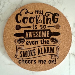 May include: A round cork trivet with a black design that says "My cooking is so awesome even the smoke alarm cheers me on!"