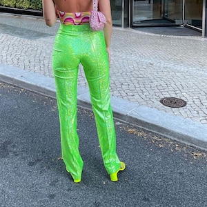 May include: A person wearing a patterned halter top, bright green sequined trousers, and neon yellow sandals. A small pink handbag is worn over the shoulder. The outfit is worn in front of a building with glass doors and a paved walkway.