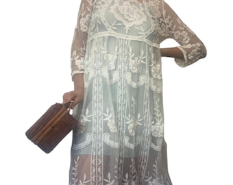 May include: A knee-length dress featuring a sheer white overlay with intricate floral embroidery and long sleeves. The dress is worn over a solid light blue slip. A small, rectangular brown woven handbag with a top handle is held at the side.