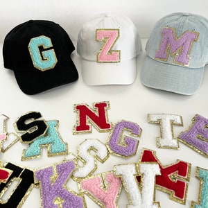 May include: A variety of colorful felt letter patches with gold trim. The patches are arranged on a white surface. There are three baseball caps with felt letter patches on them. The caps are black, white, and light blue.