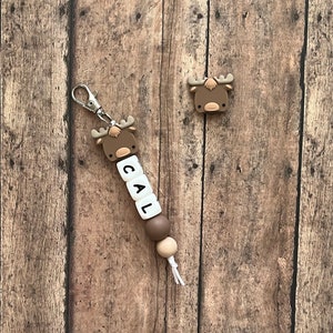 May include: A brown moose-shaped silicone keychain with the name "CAL" spelled out on white beads. The keychain has a brown and beige bead and a white string.