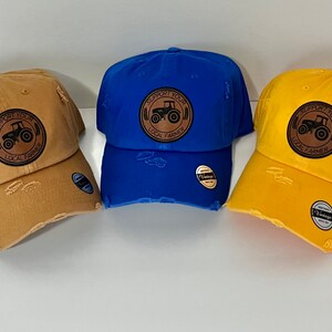 May include: Three distressed baseball caps with a brown leather patch featuring a tractor and the text "Support Your Local Farmer". The caps are in blue, yellow, and brown.