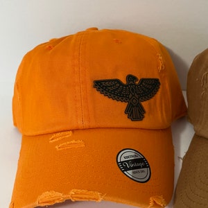 May include: Orange distressed baseball cap with a brown eagle patch. The cap has a curved brim and a vintage-style label. The fabric has a worn look with distressed edges.