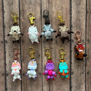 May include: A collection of whimsical keychains featuring various animal and creature designs. The keychains include cats, unicorns, and monsters, each with unique colors and textures, attached to gold or rose-gold colored clasps.