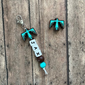 May include: A turquoise and brown beaded keychain with a turquoise bird charm and the name "JAX" spelled out on white beads. The keychain has a silver clip.