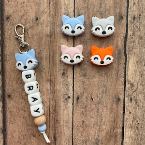 May include: A light blue fox-shaped keychain with the name "BRAY" spelled out in white lettered beads. Four additional fox-shaped charms in blue, gray, pink, and orange are also present. The items are arranged on a wooden surface.