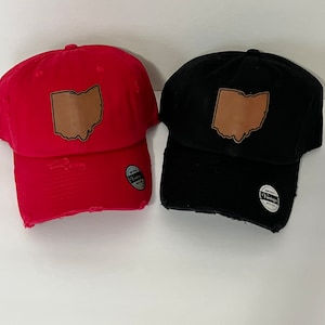 May include: Two distressed baseball caps with a brown leather patch shaped like the state of Ohio. One cap is red and the other is black.