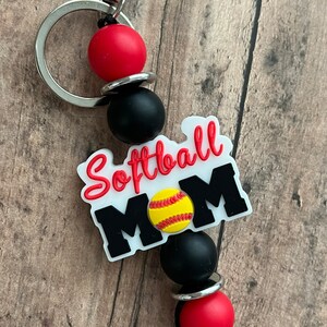 May include: A white keychain with the text "Softball Mom" and a yellow softball graphic. The keychain has black and red beads and a silver metal ring.