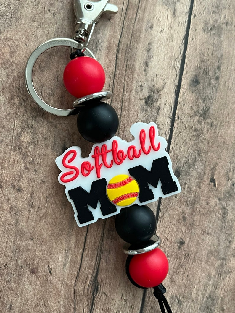 May include: A white keychain with the text "Softball Mom" and a yellow softball graphic. The keychain has black and red beads and a silver metal ring.