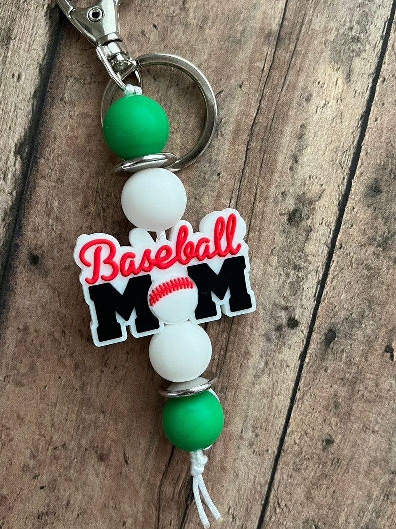 May include: A green, white, and black baseball mom keychain with a baseball graphic. The keychain has a silver ring and a white string.