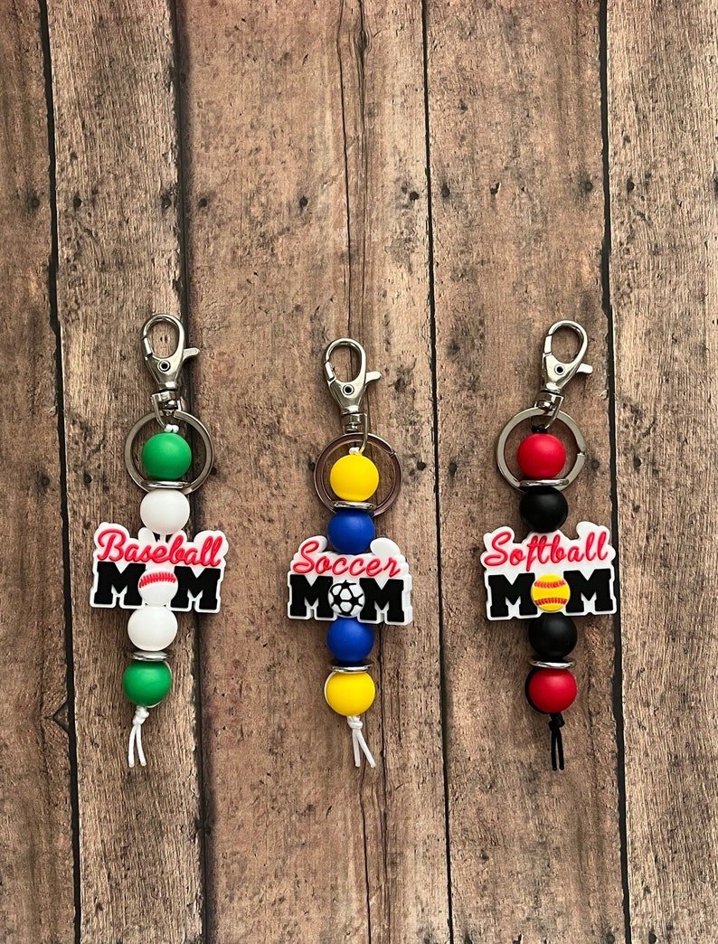 May include: Three keychains with colorful beads and charms. Each keychain features a white charm with the word "MOM" and a sports-themed graphic. The charms read "Baseball MOM", "Soccer MOM", and "Softball MOM".