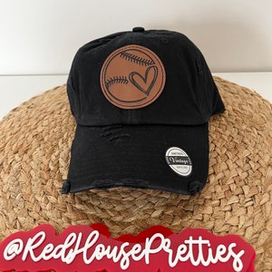 May include: Black distressed baseball cap with a brown leather patch featuring a baseball with a heart in the middle. The patch has the text "Vintage Since 2001". The cap is sitting on a woven straw surface. The text "@RedHousePretties" is visible in red letters on the surface.