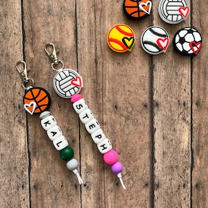 May include: Two personalized sports-themed keychains with beads and charms. One keychain has a basketball charm, the name "KAL" spelled out in white beads, and green, gray, and white beads. The other keychain has a volleyball charm, the name "STEPH" spelled out in white beads, and pink, purple, and white beads.