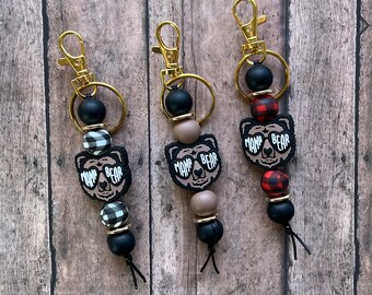 May include: Three keychains with black and white checkered, red and black checkered, and brown and black patterned beads. Each keychain features a brown bear charm with the text "Mama Bear" and black sunglasses.