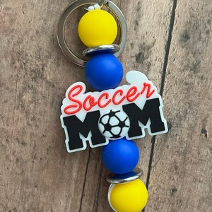 May include: A keychain with a blue and yellow silicone bead charm with a white charm that says "Soccer Mom" with a soccer ball graphic.