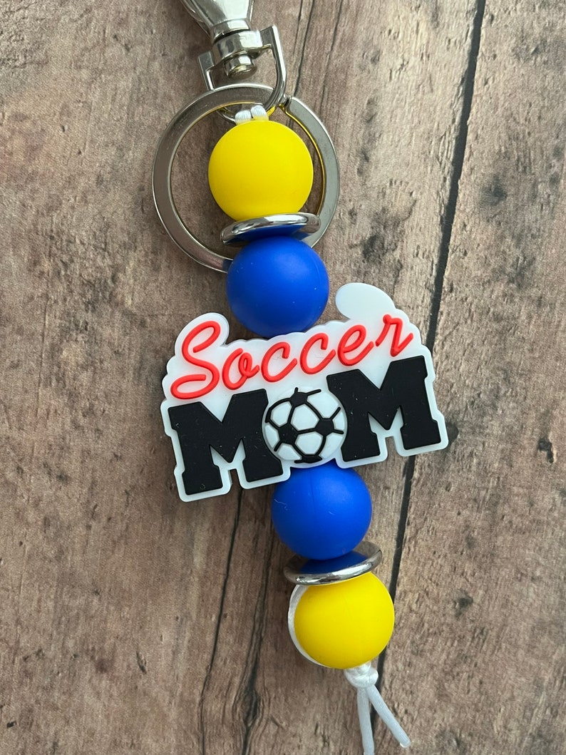 May include: A keychain with a blue and yellow silicone bead charm with a white charm that says "Soccer Mom" with a soccer ball graphic.