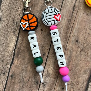 May include: Two personalized keychains with a basketball and volleyball charm, each with a small heart charm. The keychains have letter beads spelling out the names "KAL" and "STEEPH".