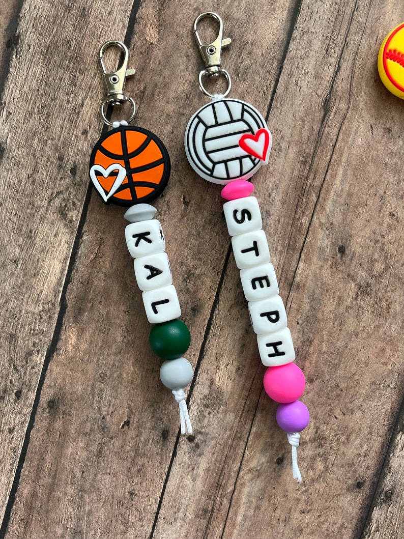 May include: Two personalized keychains with a basketball and volleyball charm, each with a small heart charm. The keychains have letter beads spelling out the names "KAL" and "STEEPH".