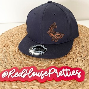 May include: A navy blue baseball cap with a brown leather catfish design. The cap has a black and white sticker on the underside of the brim that says "Premium Headwear" and "Ethos Snapback".