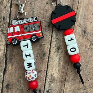May include: A red fire truck keychain with a black and red fire department badge keychain. The fire truck keychain has a white bead with the letters "W", "I", "T", and "H" on it. The fire department badge keychain has a white bead with the number "10" on it.