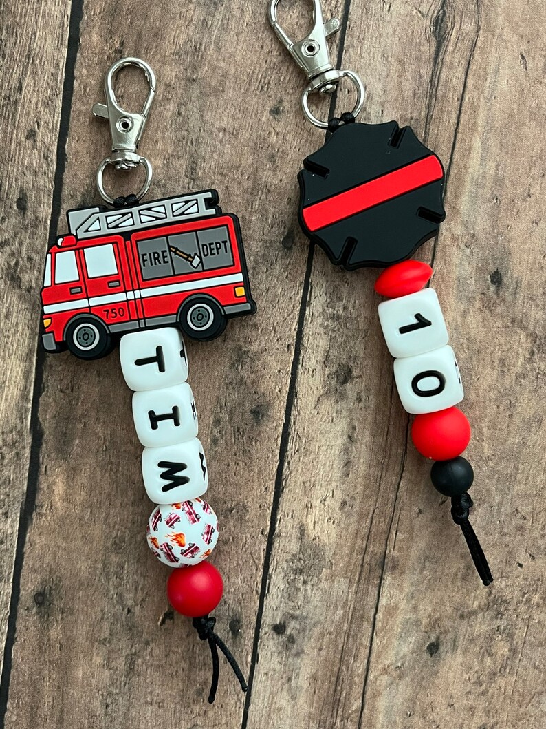 May include: A red fire truck keychain with a black and red fire department badge keychain. The fire truck keychain has a white bead with the letters "W", "I", "T", and "H" on it. The fire department badge keychain has a white bead with the number "10" on it.