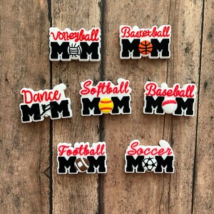 May include: Seven different sports-themed "Mom" shoe charms. Each charm features a different sport, including volleyball, basketball, dance, softball, baseball, football, and soccer. The charms are all white with black lettering and red accents. Each charm features a small graphic of the sport's ball.