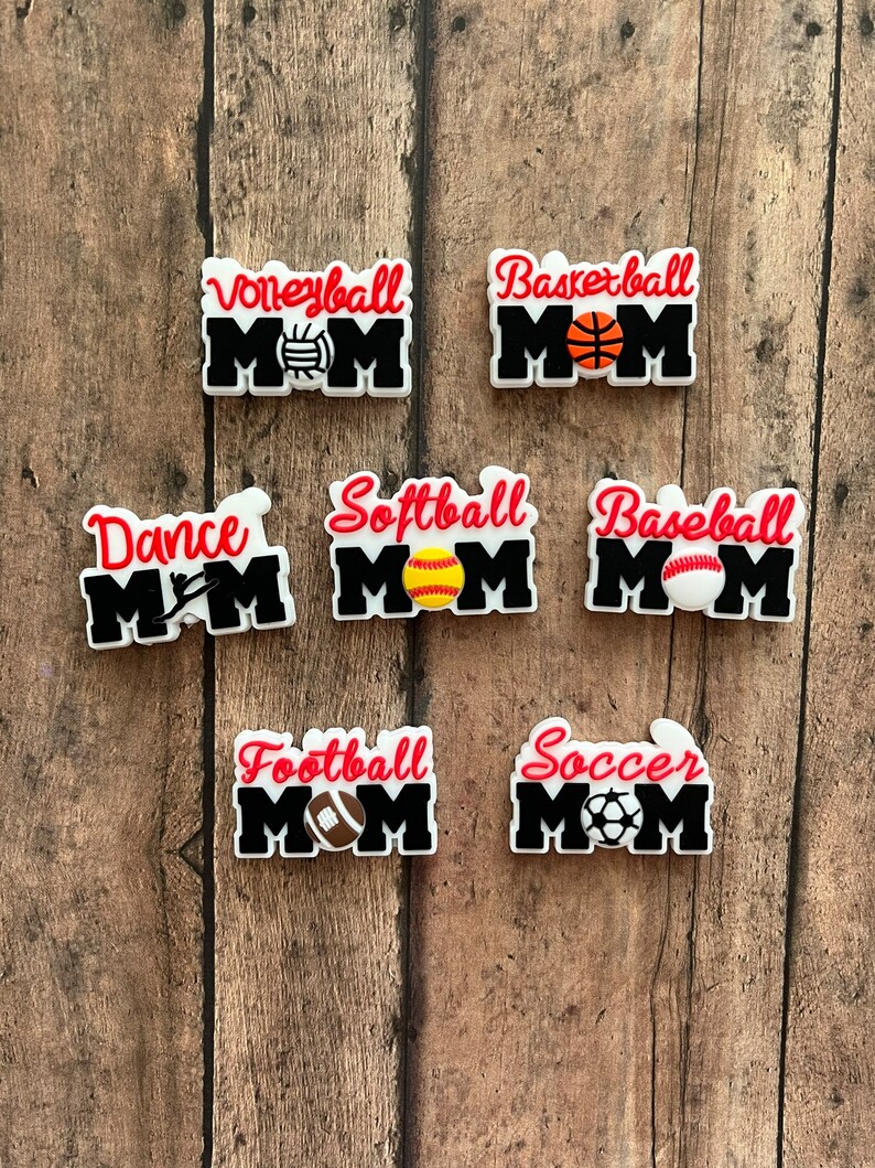 May include: Seven different sports-themed "Mom" shoe charms. Each charm features a different sport, including volleyball, basketball, dance, softball, baseball, football, and soccer. The charms are all white with black lettering and red accents. Each charm features a small graphic of the sport's ball.