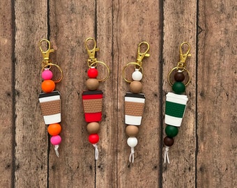 May include: Four coffee cup keychains with colorful beads. Each keychain has a different color scheme: pink and orange, red and brown, white and brown, and green and brown.