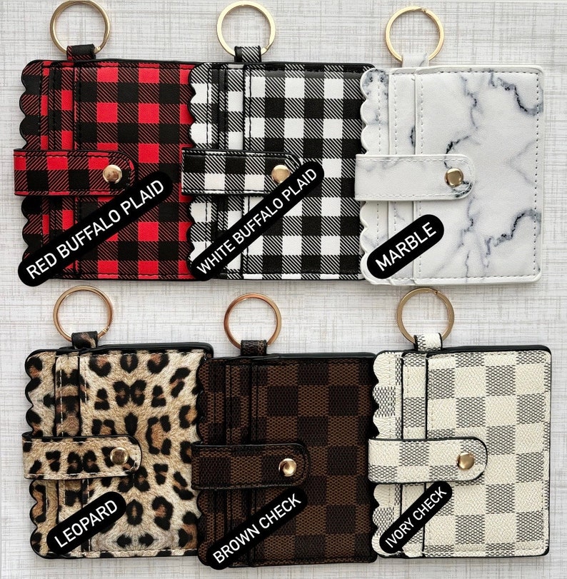May include: Six different keychains with card holders. The keychains are in various patterns including red buffalo plaid, white buffalo plaid, marble, leopard, brown check, and ivory check.