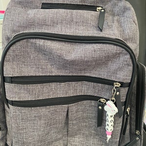 May include: A gray backpack with black accents and a zippered pocket. A white beaded keychain with the word "ROXY" is attached to the zipper pull.