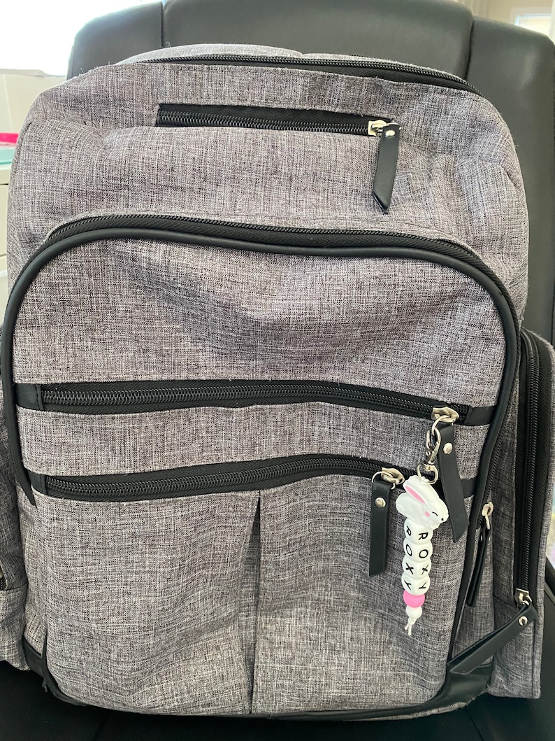 May include: A gray backpack with black accents and a zippered pocket. A white beaded keychain with the word "ROXY" is attached to the zipper pull.
