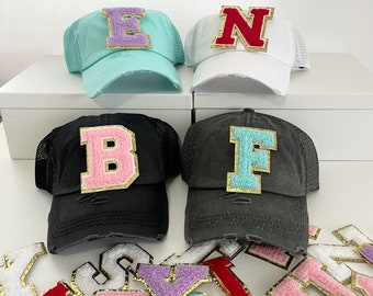 May include: Four trucker hats with different colored letters on the front. The letters are "E", "N", "B", and "F". The hats are in various colors, including black, white, and light blue. There are also several letter patches in different colors and styles.