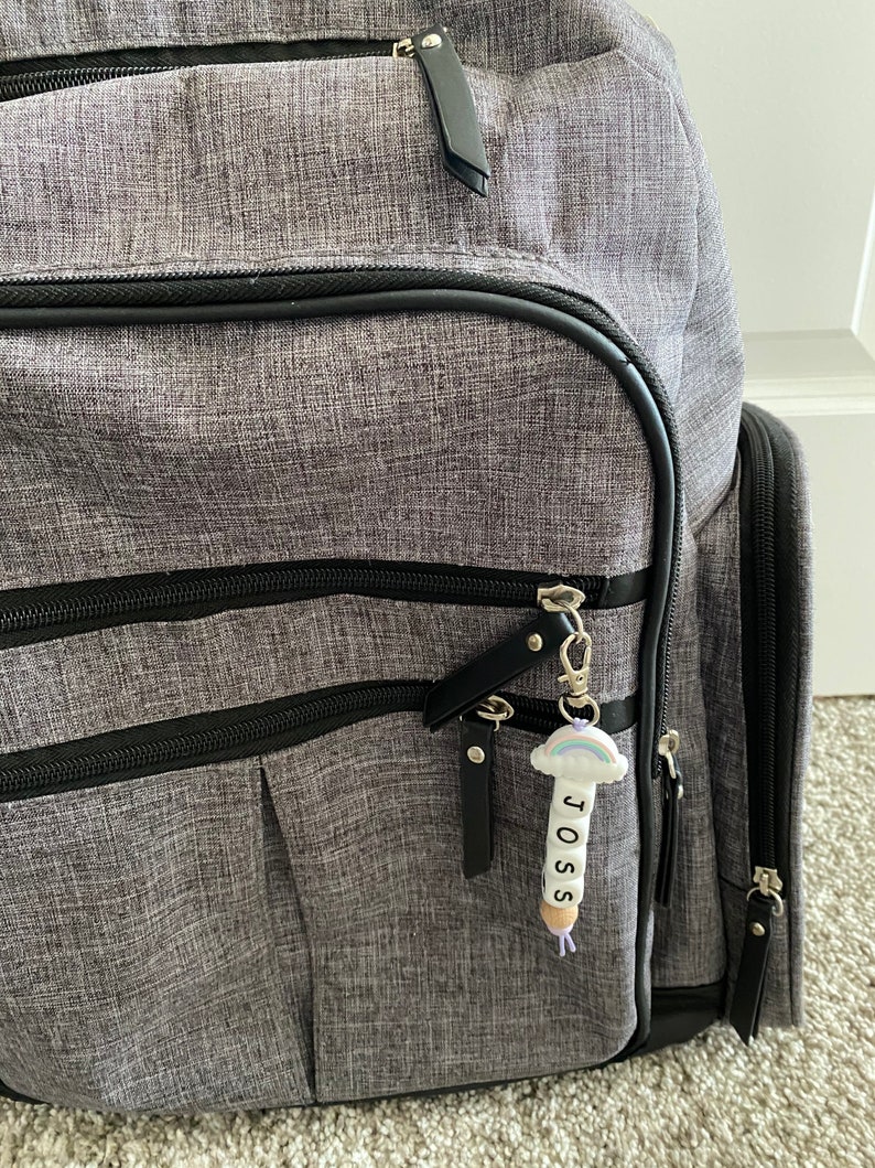 May include: A gray backpack with black accents and a zipper pull with a rainbow charm and the name "JOSS" spelled out on it.
