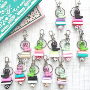 May include: A collection of keychains featuring colorful worm-like figures wearing glasses, perched atop miniature stacks of books. Each keychain has a silver clasp and a decorative bead at the bottom. The worms are in various colors, including pink, green, and black.