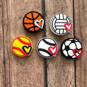 May include: Six colorful sports ball charms with a heart design. The charms are made of silicone and feature a basketball, volleyball, softball, baseball, and soccer ball.