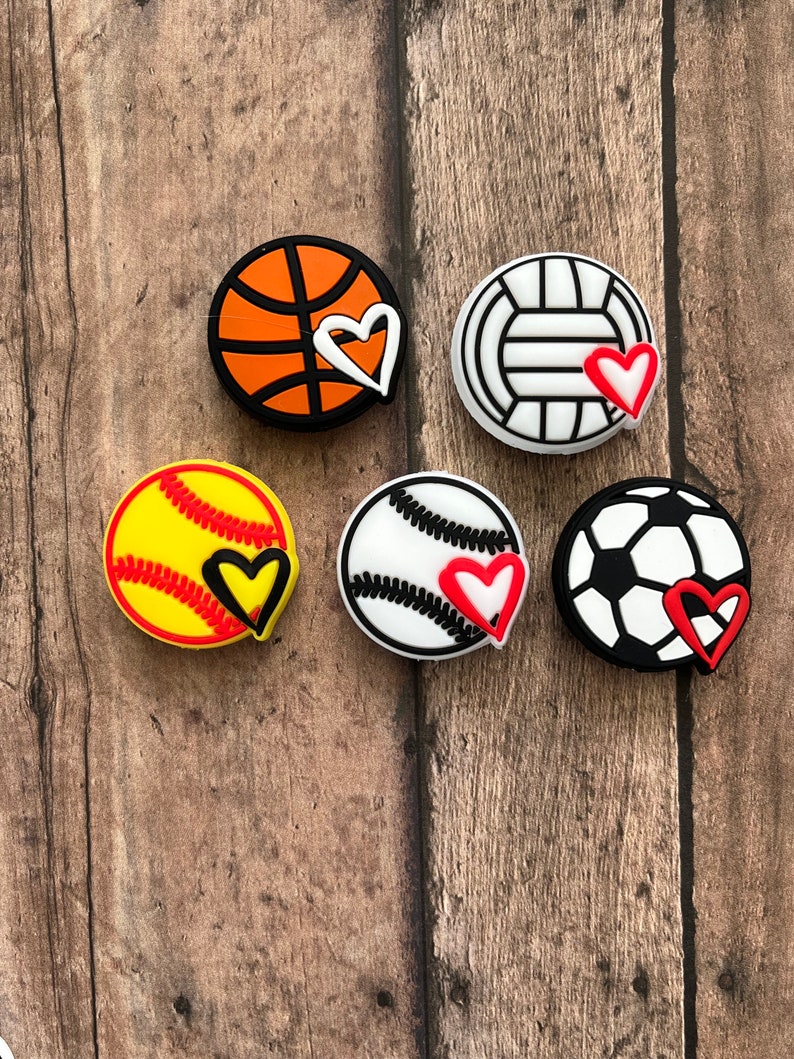 May include: Six colorful sports ball charms with a heart design. The charms are made of silicone and feature a basketball, volleyball, softball, baseball, and soccer ball.