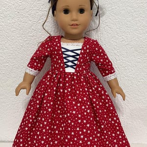 May include: A doll wearing a red dress with white star patterns. The dress has a white lace-trimmed neckline and sleeves, and a blue ribbon corset detail. The doll has brown hair styled in a bun with a blue ribbon.