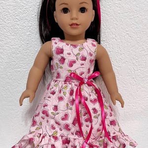May include: A doll wearing a pink dress with a heart and leaf pattern. The dress has a pink ribbon tied around the waist and a ruffled hem. The doll also wears pink sandals and a pink headband. The doll has black hair and brown eyes.