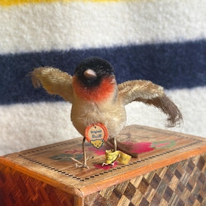 May include: A vintage Steiff bird plush toy with outstretched wings, featuring a black head, red chest, and tan body. The toy has a Steiff tag and is perched on a wooden box with a geometric design.